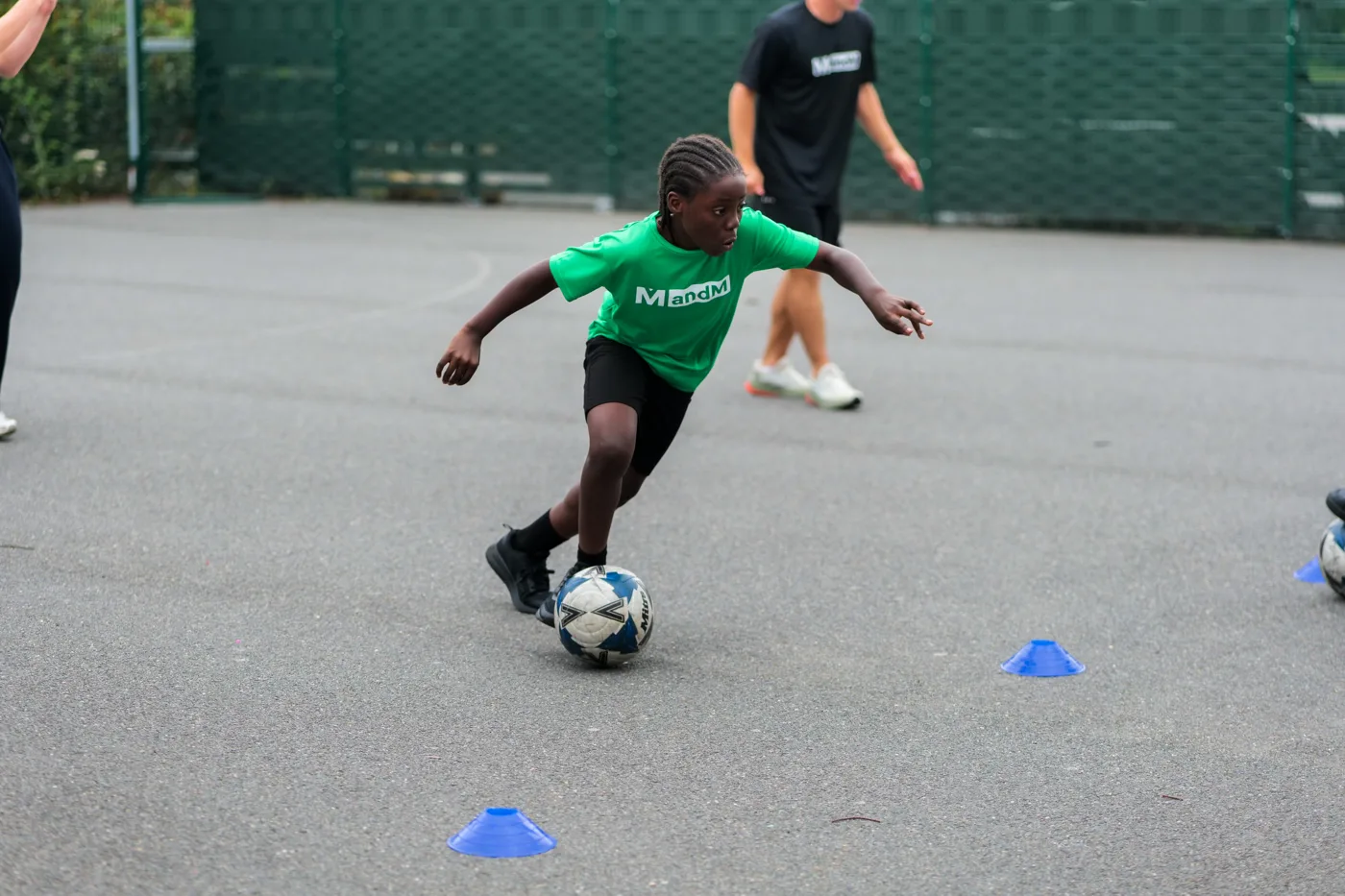 A pupil in a green MandM shirt dribbling a football between cones on the playground