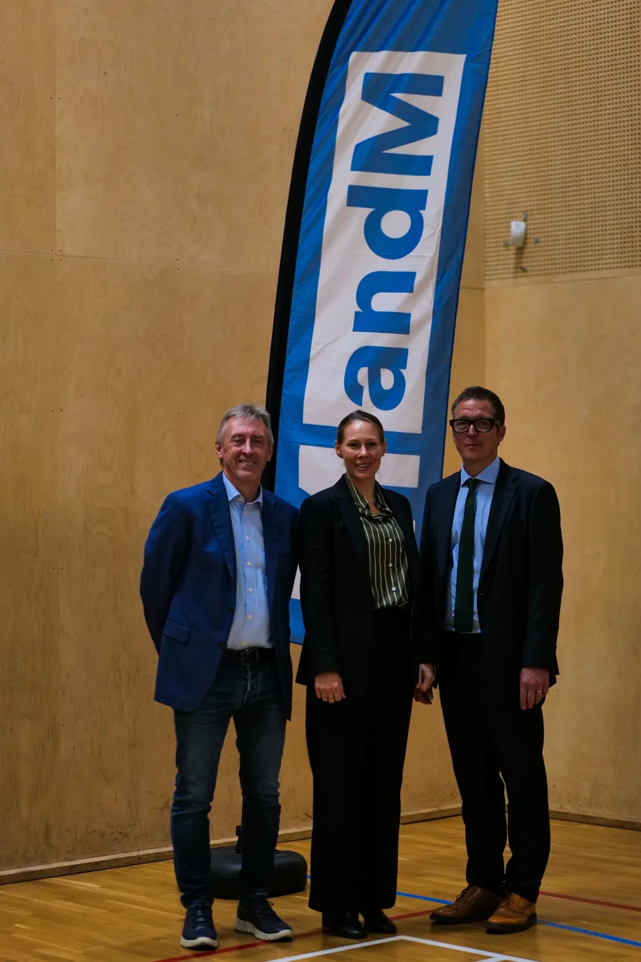 Two men in suits and a woman standing in front of an MandM banner in the sports hall