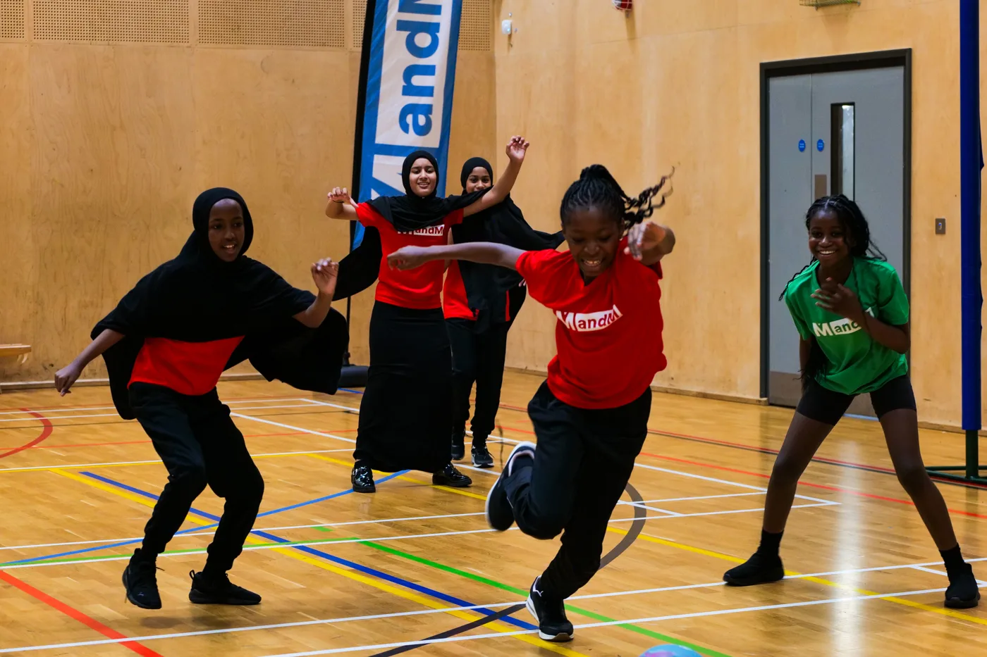 Pupils in red and green shirts, some wearing hijabs, running for the ball in the sports hall