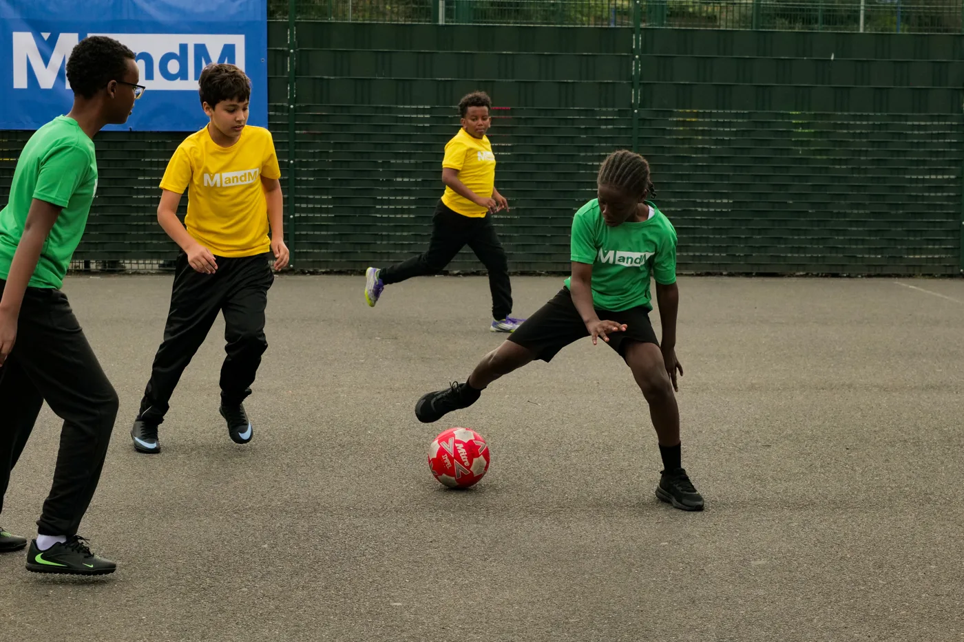 Pupils in green and yellow MandM shirts chasing a red football on the outdoor court