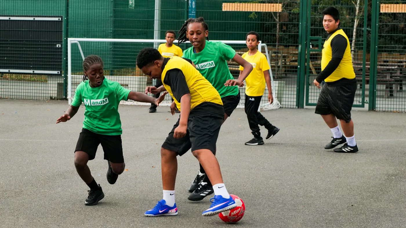 A pupil in a yellow bib shielding the ball from a pupil in a green shirt in front of a goal