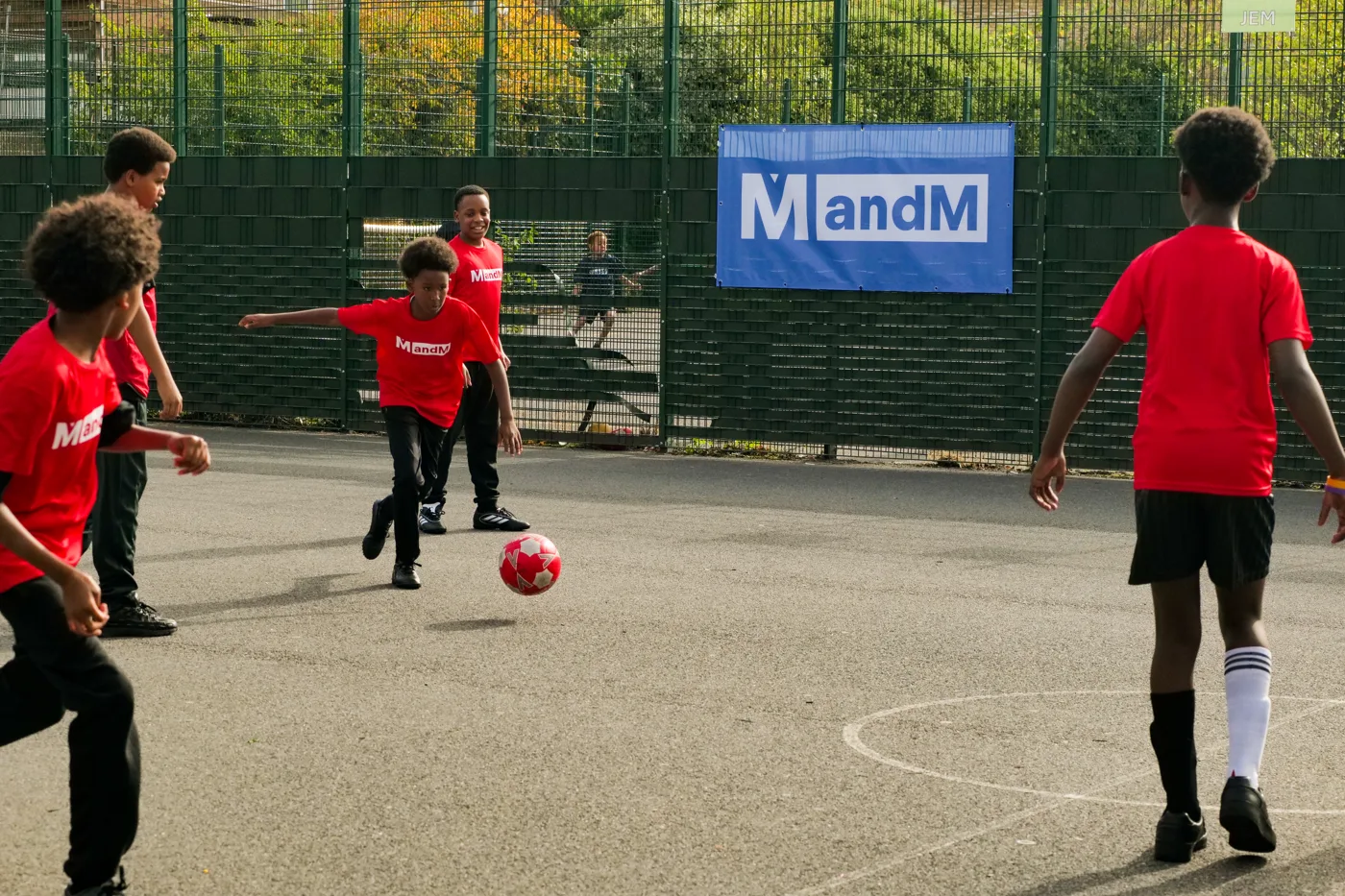 Pupils in red MandM shirts playing football on the outdoor court beside an MandM banner