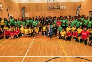 Harris Academy Clapham pupils in red, yellow and green shirts sitting and standing in the sports hall with Mike Tomkins, Harry Cain and Beth Durant