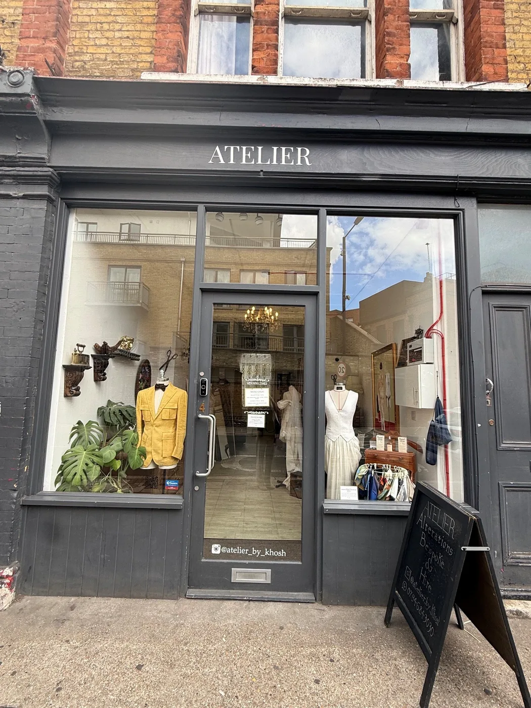 The shopfront of Atelier on Landor Road, a dark grey frontage with a tailor's window display and an A-board reading alterations, bespoke and hire