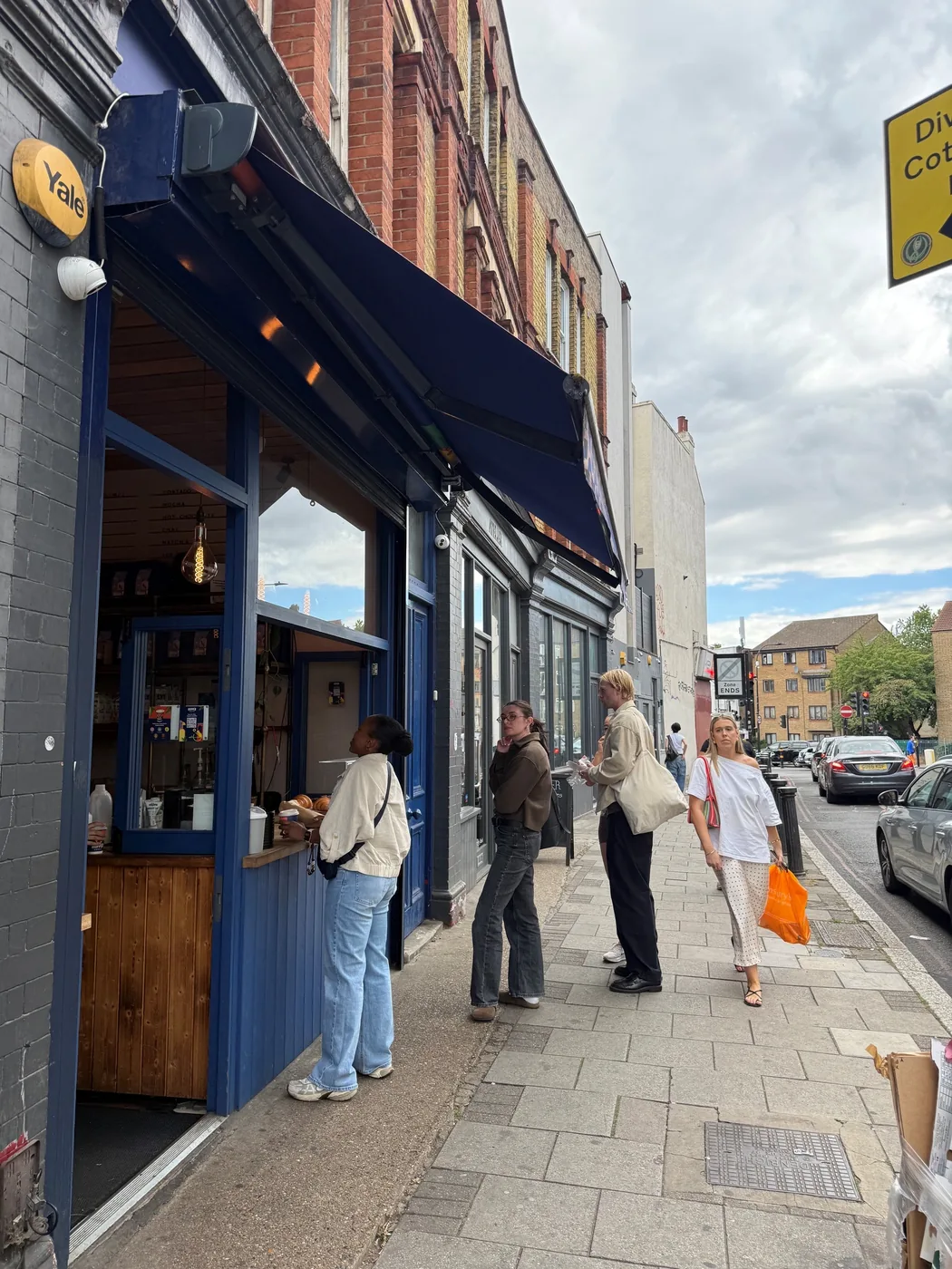 People standing at a street-facing coffee hatch on Landor Road, with parked cars and a diversion sign further down the street